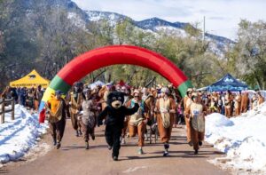 Dozens pf people wearing bear suits run in a forward direction led by a person wearing a bear mascot suit with colorful red arch and snow covered mountains in background