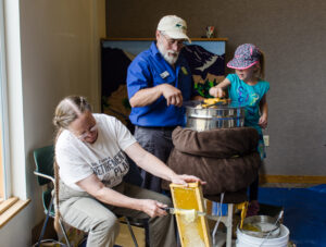 Person scrapes honey from frame while an adult assists a child in using honey extractor machine