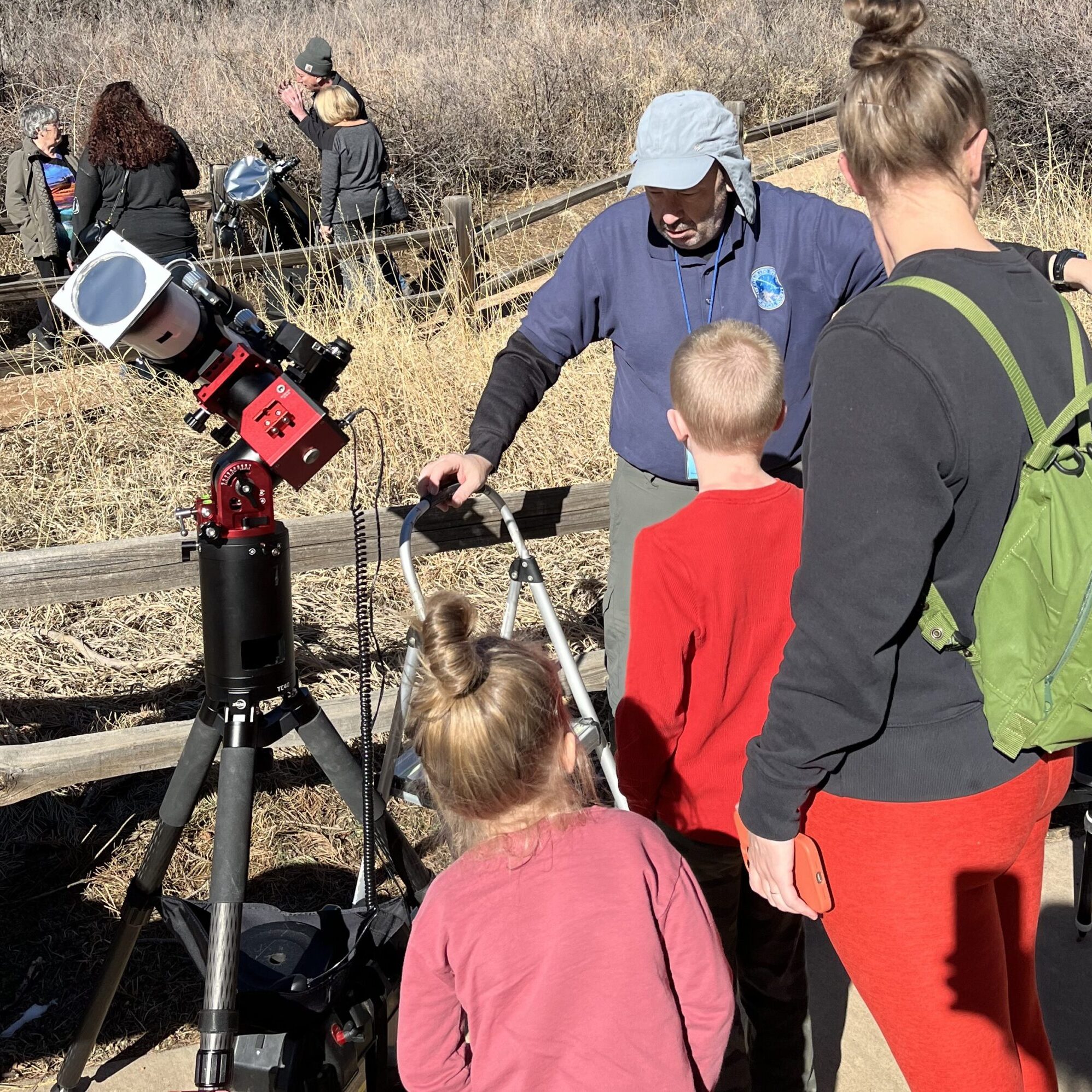 Sidewalk Astronomy Adult with 2 children stands near telescope operated by a man wearing a sun hat