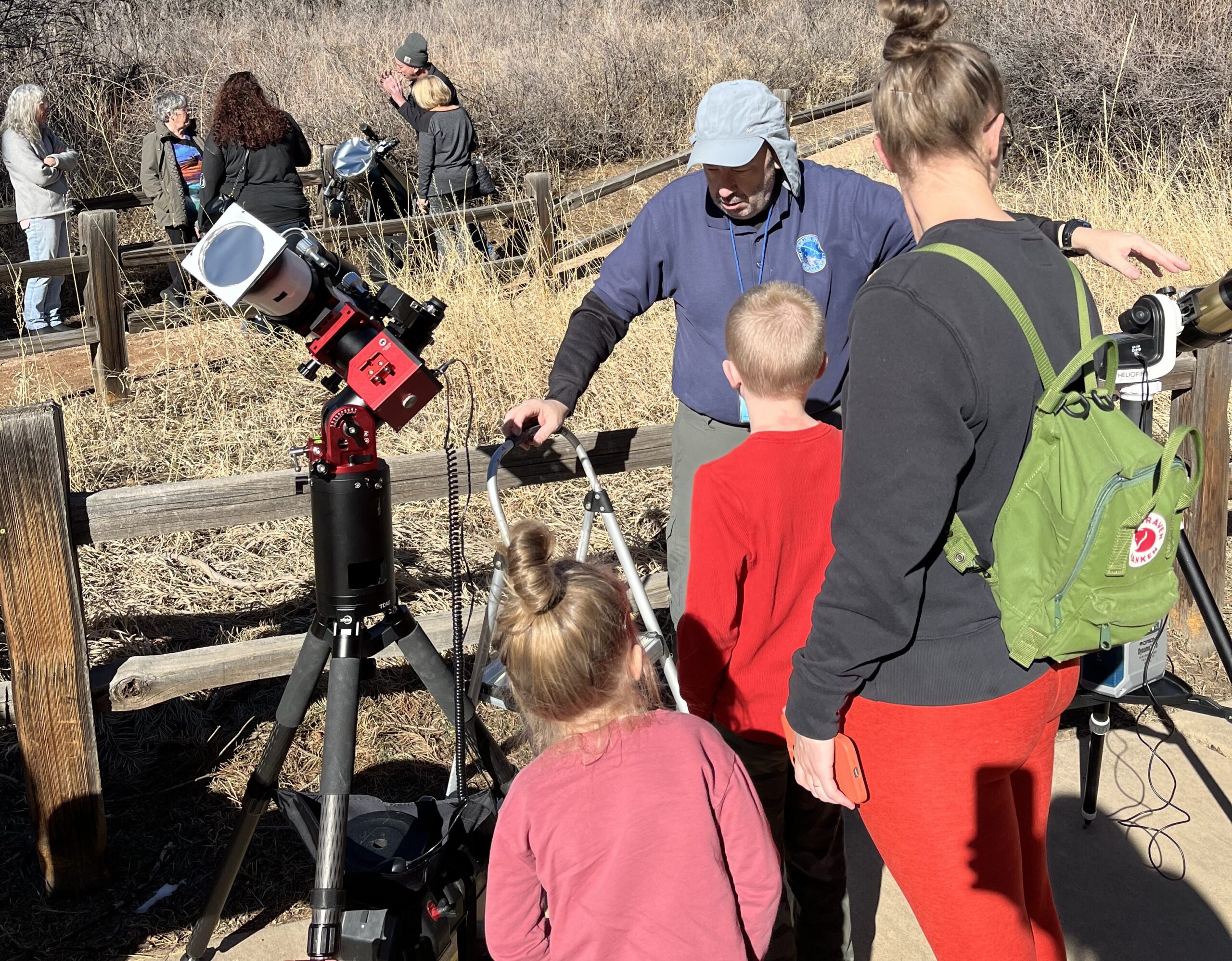 Sidewalk Astronomy Adult with 2 children stands near telescope operated by a man wearing a sun hat