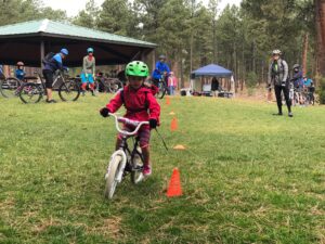Young child wearing green helmet and red jacket rides bike beside an orange cone in foreground. In background, there are about ten people standing or riding bikes.
