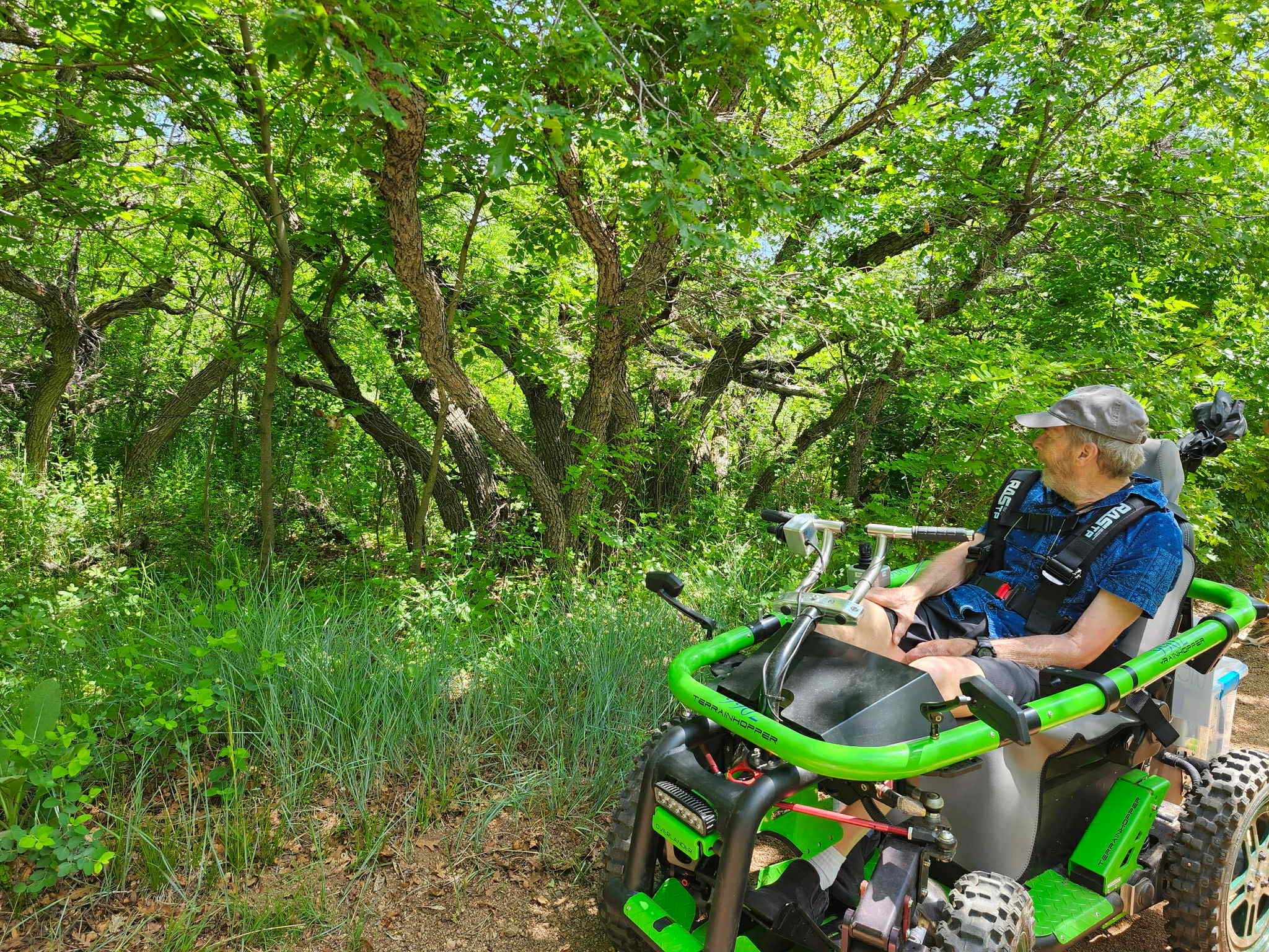 A man sits in a green, off-road mobility device and looks away from the camera at green grasses and trees.