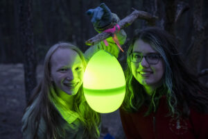 Two young people smile in the dark with a large glowing easter egg between them.