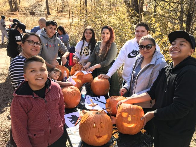 A family smiles as they carve pumpkins into jack-o-lanterns outside on a fall day.