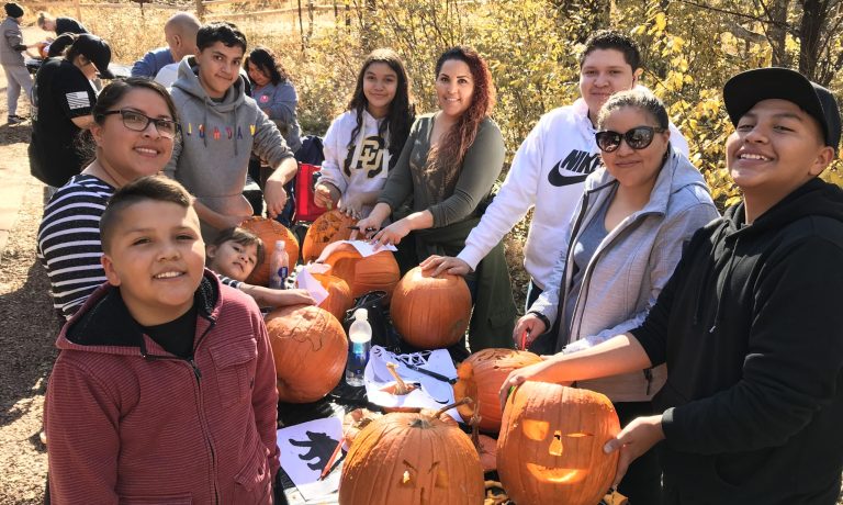 A family smiles as they carve pumpkins into jack-o-lanterns outside on a fall day.