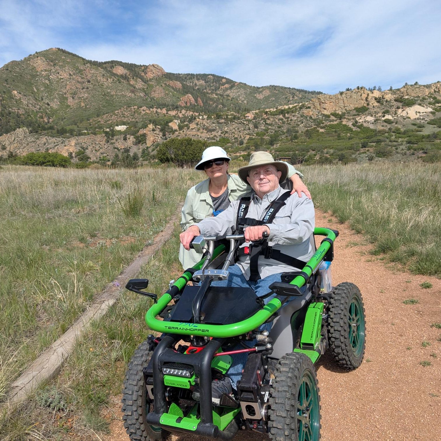 BCNC Trailability Participant and Companion In a meadow at the base of some foothills mountains, a man holds the handlebars to a green, off-road mobility device while his companion wraps and arm around him and they smile at he camera.