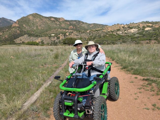 BCNC Trailability Participant and Companion In a meadow at the base of some foothills mountains, a man holds the handlebars to a green, off-road mobility device while his companion wraps and arm around him and they smile at he camera.