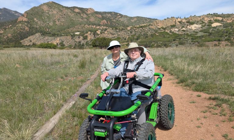 BCNC Trailability Participant and Companion In a meadow at the base of some foothills mountains, a man holds the handlebars to a green, off-road mobility device while his companion wraps and arm around him and they smile at he camera.