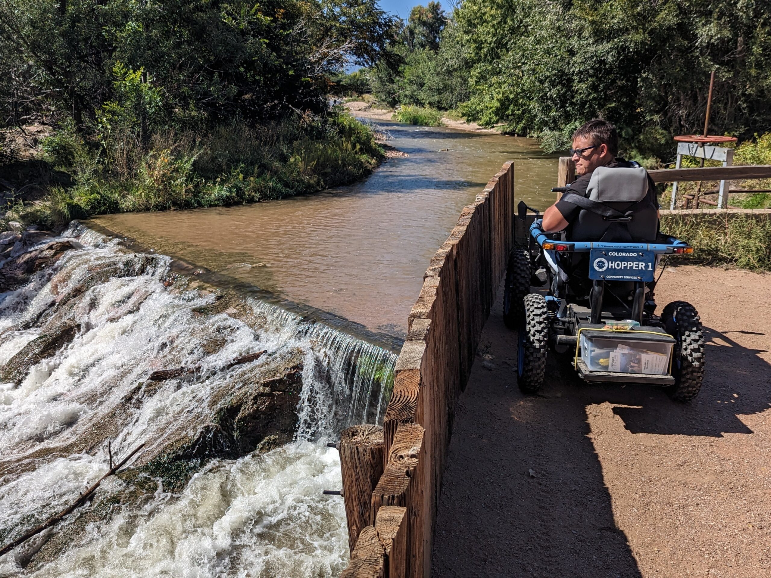 A man sits in a blue, 0ff-road mobility device while looking over a wooden fence at a rocky waterfall.